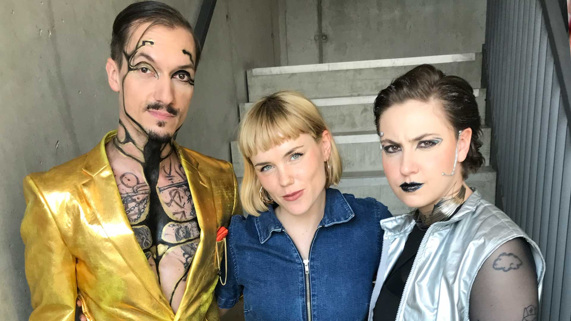 Ein Mann futuristisch geschminkter Mann mit goldener Jacke, eine blonde Frau mit Jeansoverall und eine dunklhaarige Frau mit futuristischer Schminke und einer silbernen Weste stehen in einem Treppenaufgang und schauen in die Kamera.  (Foto: SWR)